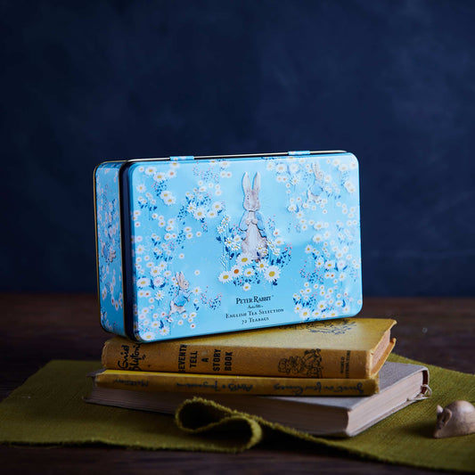 A rectangular blue metal tin with a Peter Rabbit floral print sits on top of stacked vintage-style books on a green cloth, against a dark blue background.
