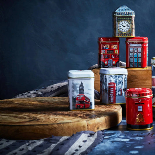 A stack of London-themed metal tins stacked on a wooded bread board and box, featuring Big Ben, red double-decker buses, red telephone booths, and a red post box design.