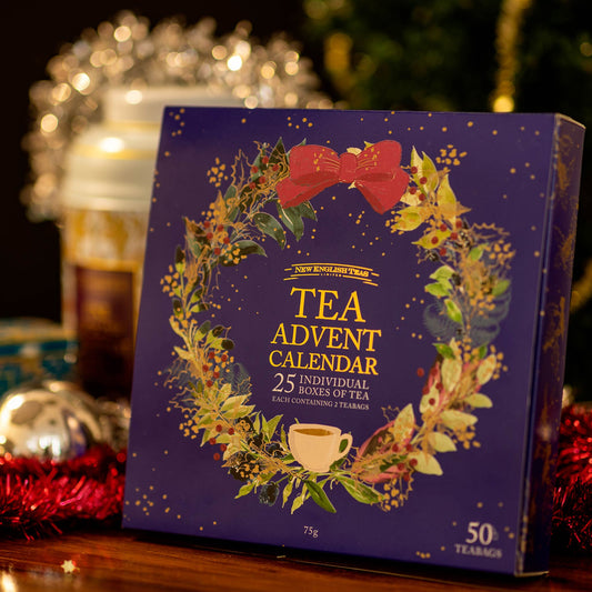 A square tea advent calendar gift box standing upright on a wooden surface. The box is dark blue with a decorative wreath made of green leaves, red berries, gold accents, and a red bow at the top, plus a small teacup illustration near the bottom.