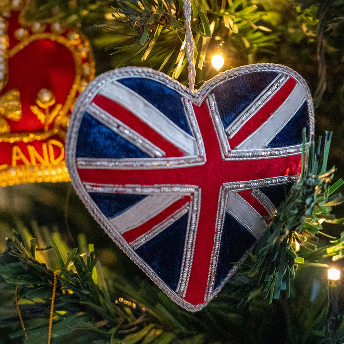 A heart-shaped Union Jack Christmas ornament hanging on a green Christmas tree with warm string lights.