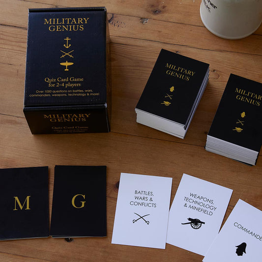 MILITARY GENIUS quiz card game set arranged on a wooden tabletop, including the product box, two deck stacks, and several cards. The box and cards are black with gold lettering and symbols, while some cards are white with black text and icons.