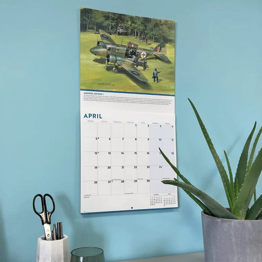 A vertical wall calendar featuring a vintage military aircraft, an Airspeed Oxford 1, illustration at the top and a monthly calendar grid below, hanging on a light blue wall. The scene includes a potted aloe plant on the right and a small holder with scissors and pens on the left.