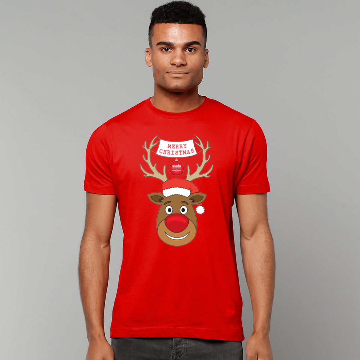 A man wearing a red short-sleeve crewneck t-shirt featuring a cartoon graphic of a reindeer with brown antlers, a red nose, a Santa hat, and a banner that reads 'MERRY CHRISTMAS'.