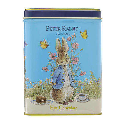 A round metal tin for Peter Rabbit Hot Chocolate featuring a light blue background with an illustration of Peter Rabbit in a blue jacket, surrounded by flowers, butterflies, a chocolate bar, and a cup of hot chocolate. The tin has a dark blue lid and gold trim, with 'PETER RABBIT' and 'Peter Rabbit' printed at the top and 'Hot Chocolate' at the bottom.