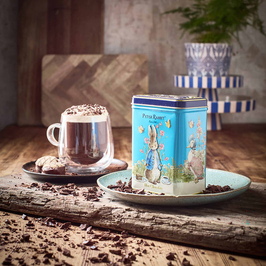 A blue Peter Rabbit tea tin with illustrated Peter Rabbit sits on a light blue plate on a rustic wooden table, alongside a glass mug of hot chocolate with whipped cream and chocolate shavings. The background features a warm, cozy interior with a striped side table and a blurred plant.