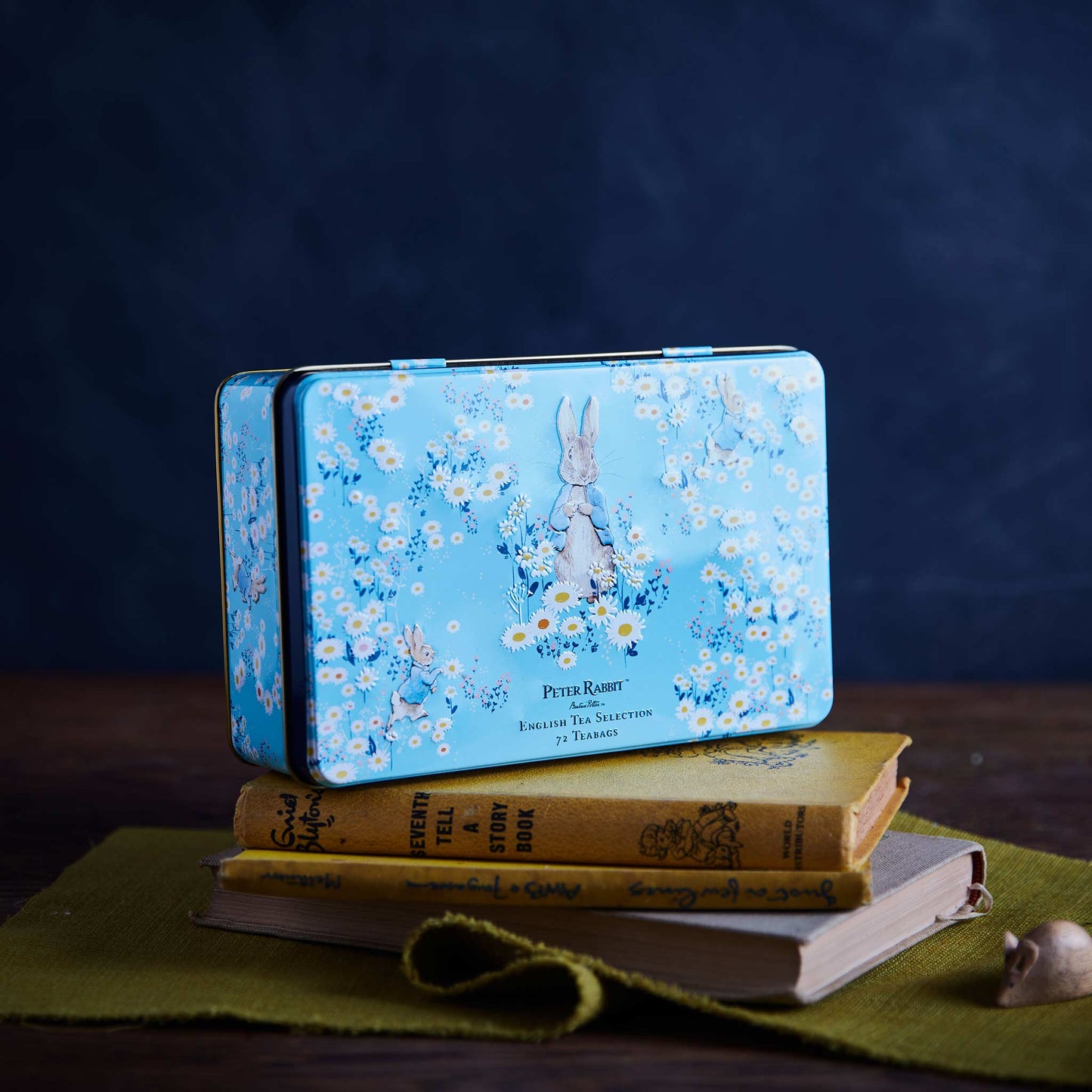 A rectangular blue metal tin with a Peter Rabbit floral print sits on top of stacked vintage-style books on a green cloth, against a dark blue background.