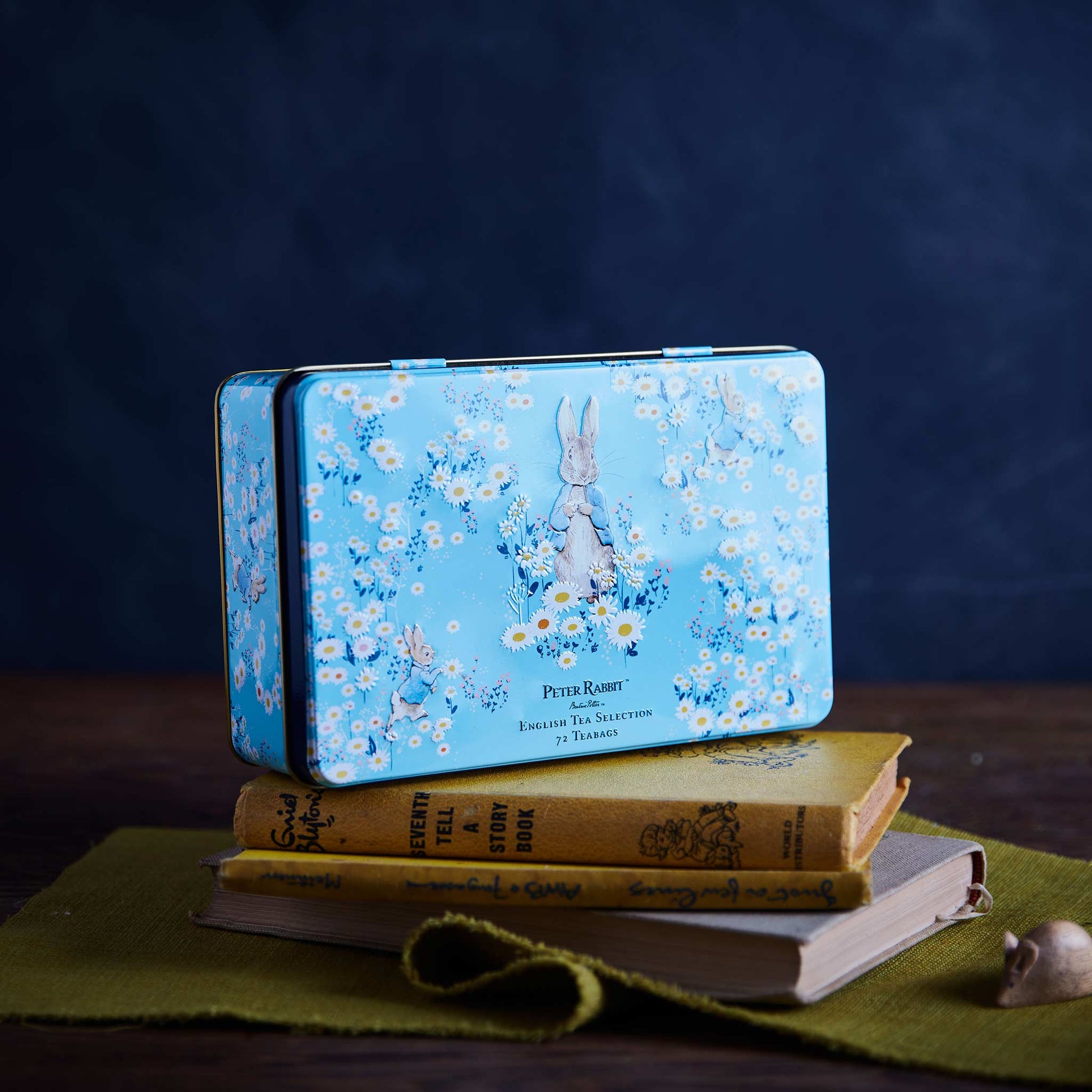 A rectangular blue metal tin with a Peter Rabbit floral print sits on top of stacked vintage-style books on a green cloth, against a dark blue background.