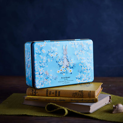 A rectangular blue metal tin with a Peter Rabbit floral print sits on top of stacked vintage-style books on a green cloth, against a dark blue background.