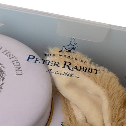 A close-up of a Peter Rabbit themed gift set inside a light blue box. Visible items include a round white tin with blue 'English' lettering, a cream-colored plush toy or accessory with 'The World of Peter Rabbit' branding, and a beige faux-fur trim.