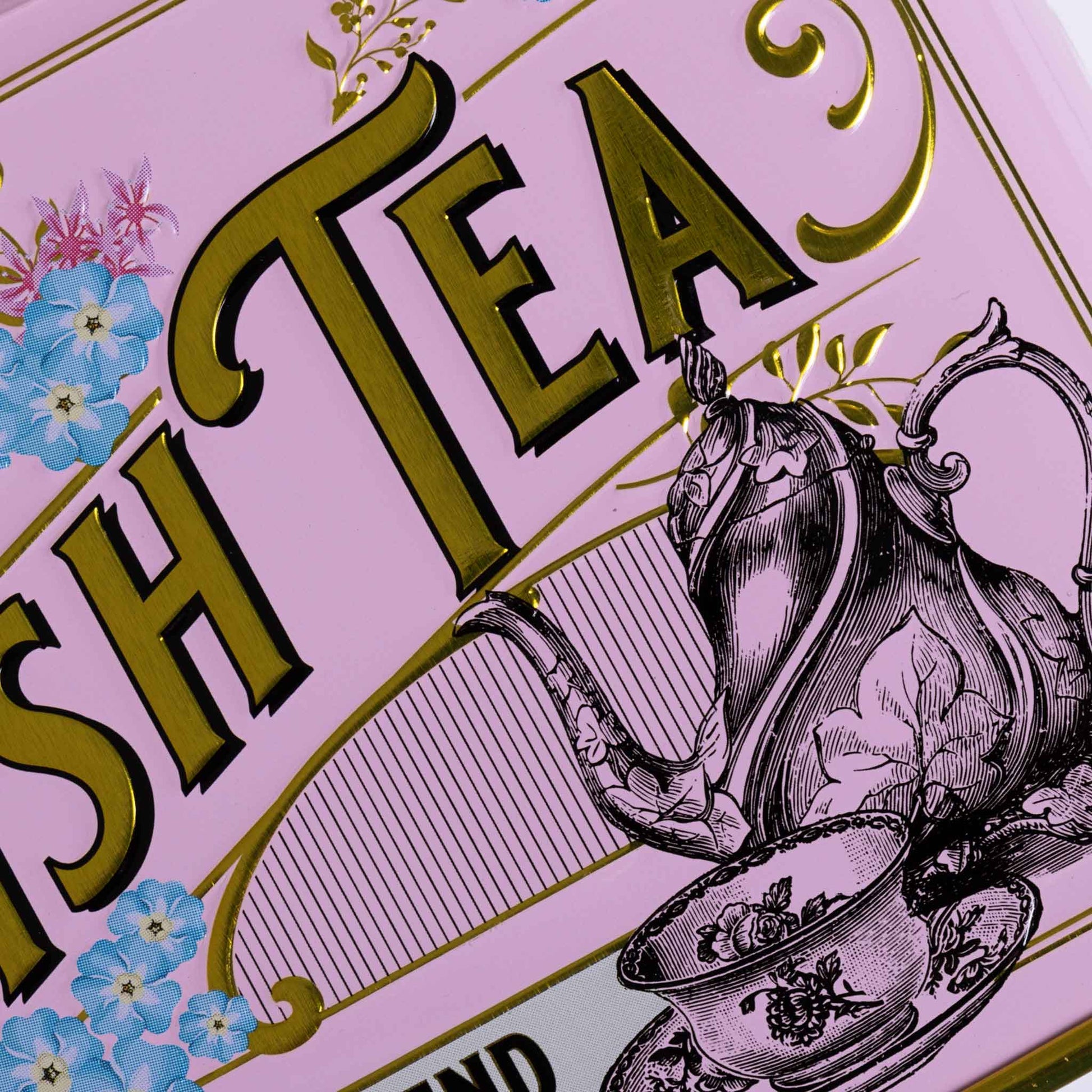 Close-up of a pink tea tin label with large gold 'TEA' lettering, blue flowers, and a black-and-white teapot and teacup illustration.