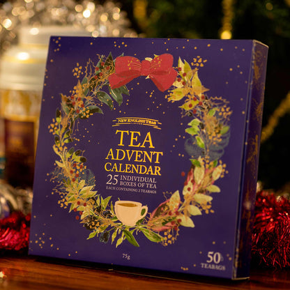 A square tea advent calendar box in deep purple with a festive wreath illustration, red bow, gold text reading 'TEA ADVENT CALENDAR,' and a small teacup graphic, photographed on a wooden surface with holiday decorations blurred in the background.