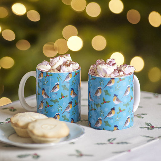 Two mugs with Military Robin pattern filled with hot chocolate and marshmallows, on a table with a blurred sparkly Christmas light festive background. On a table runner with Holly with berries design. Mug with military Robin design on a blue background. Patterned Mugs with Robins each wearing a hat of the Army, Royal Navy and RAF, with holly and mistletoe and holly on a blue background.