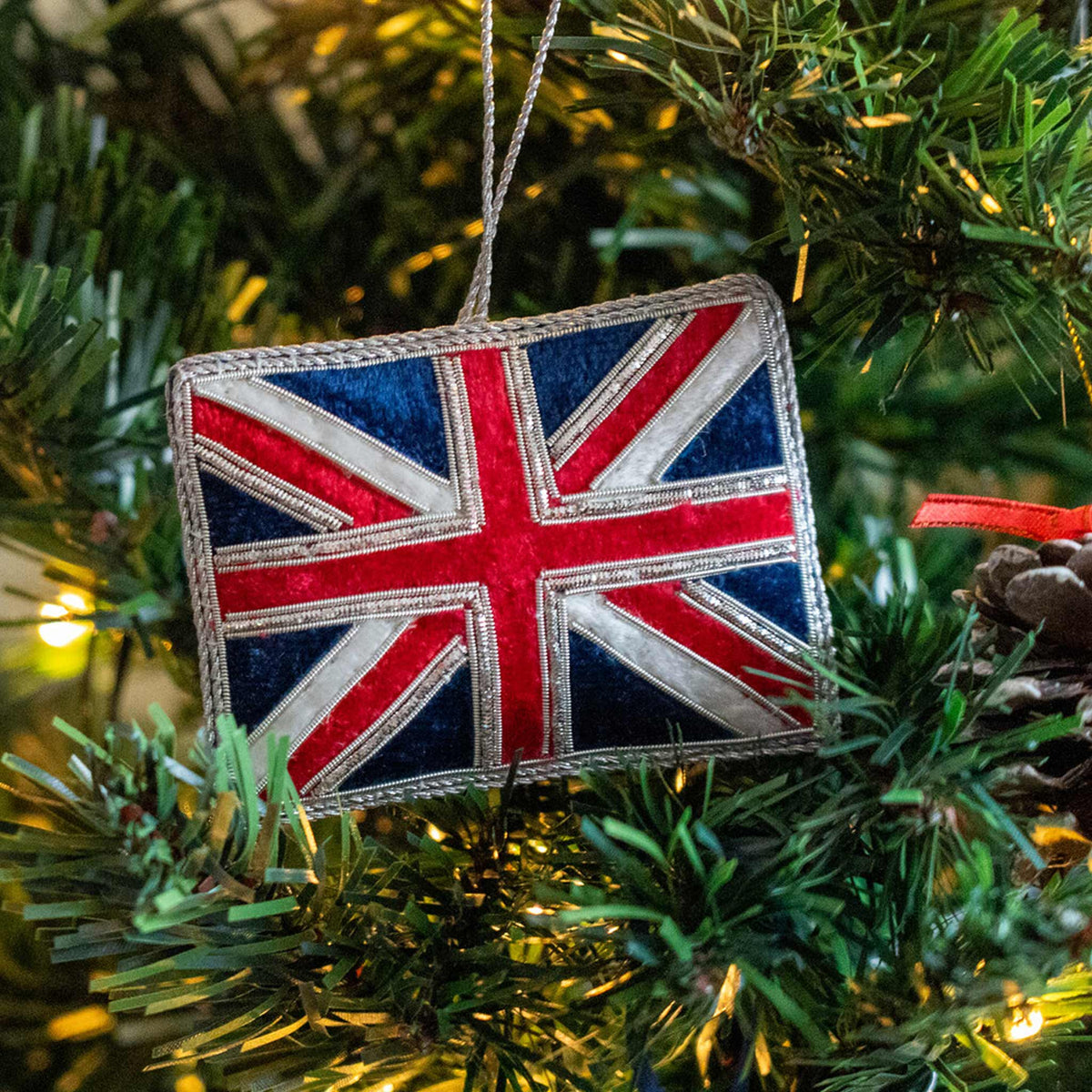 A Union Jack flag-shaped hanging ornament with a silver hanging loop, shown hanging in a Christmas Tree.