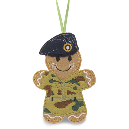 A felt gingerbread soldier Christmas ornament wearing a dark navy blue beret and Army uniform: green, brown, and tan camouflage, hanging from a light green ribbon on a white background.