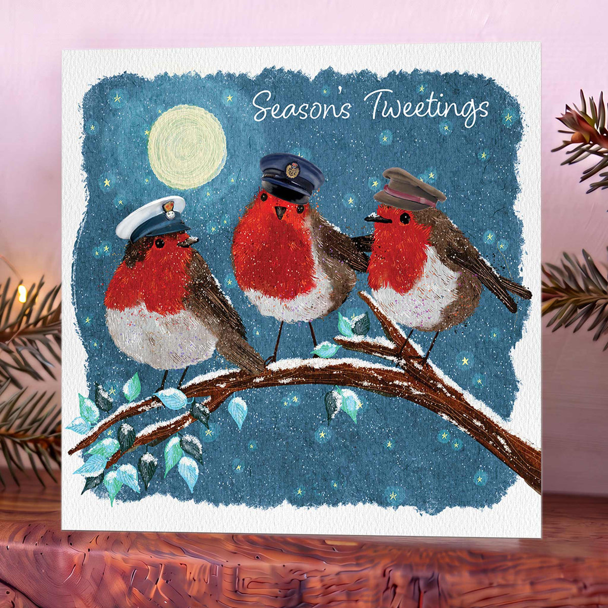 Christmas card showing three robins in Army, Royal Navy and RAF hats on a branch beneath a moonlit blue sky, with ‘Seasons Tweetings’ above. Shot on wooden mantelpiece.