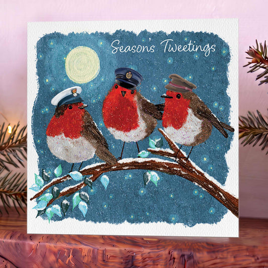 Christmas card showing three robins in Army, Royal Navy and RAF hats on a branch beneath a moonlit blue sky, with ‘Seasons Tweetings’ above. Shot on wooden mantelpiece.