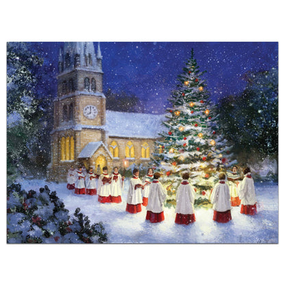 A folded greeting card standing upright on a white background, printed with a festive winter Christmas scene showing a church, snow, a decorated tree, and a choir in white robes with red skirts.