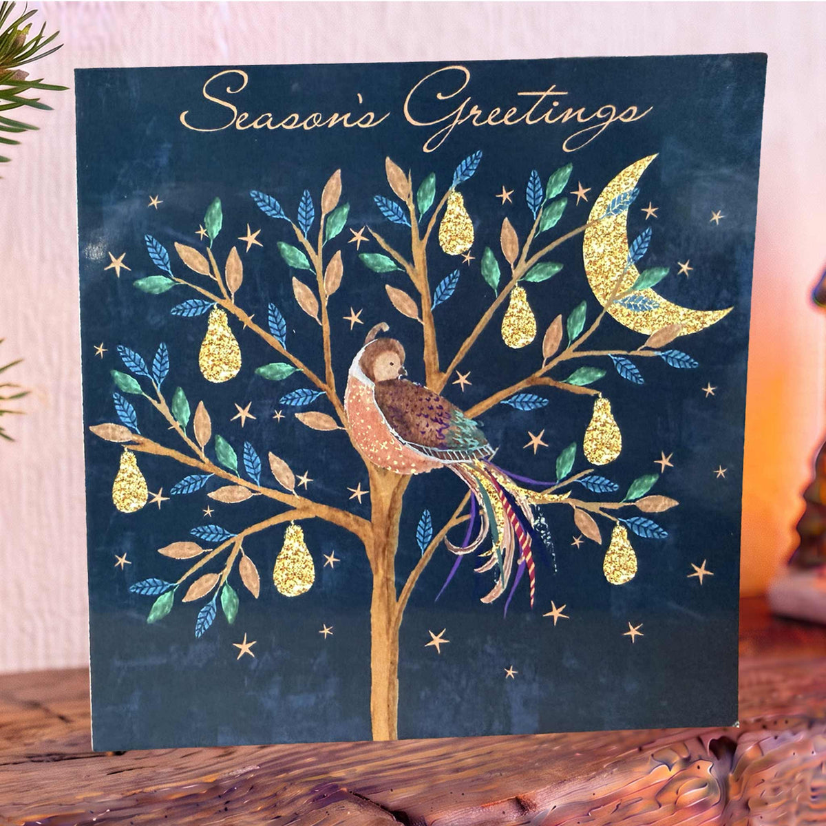 Navy Christmas Card on mantelpiece with a pear tree with gold pears Partridge, stars and crescent moon, topped with gold ‘Season’s Greetings’.