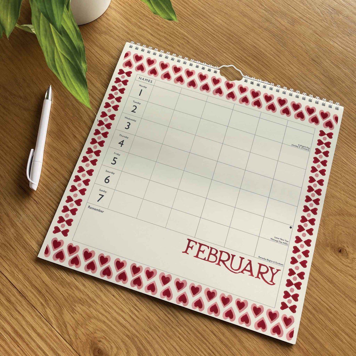 A spiral-bound February calendar page with a red heart border, shown on a wooden desk next to a white pen and a green plant.