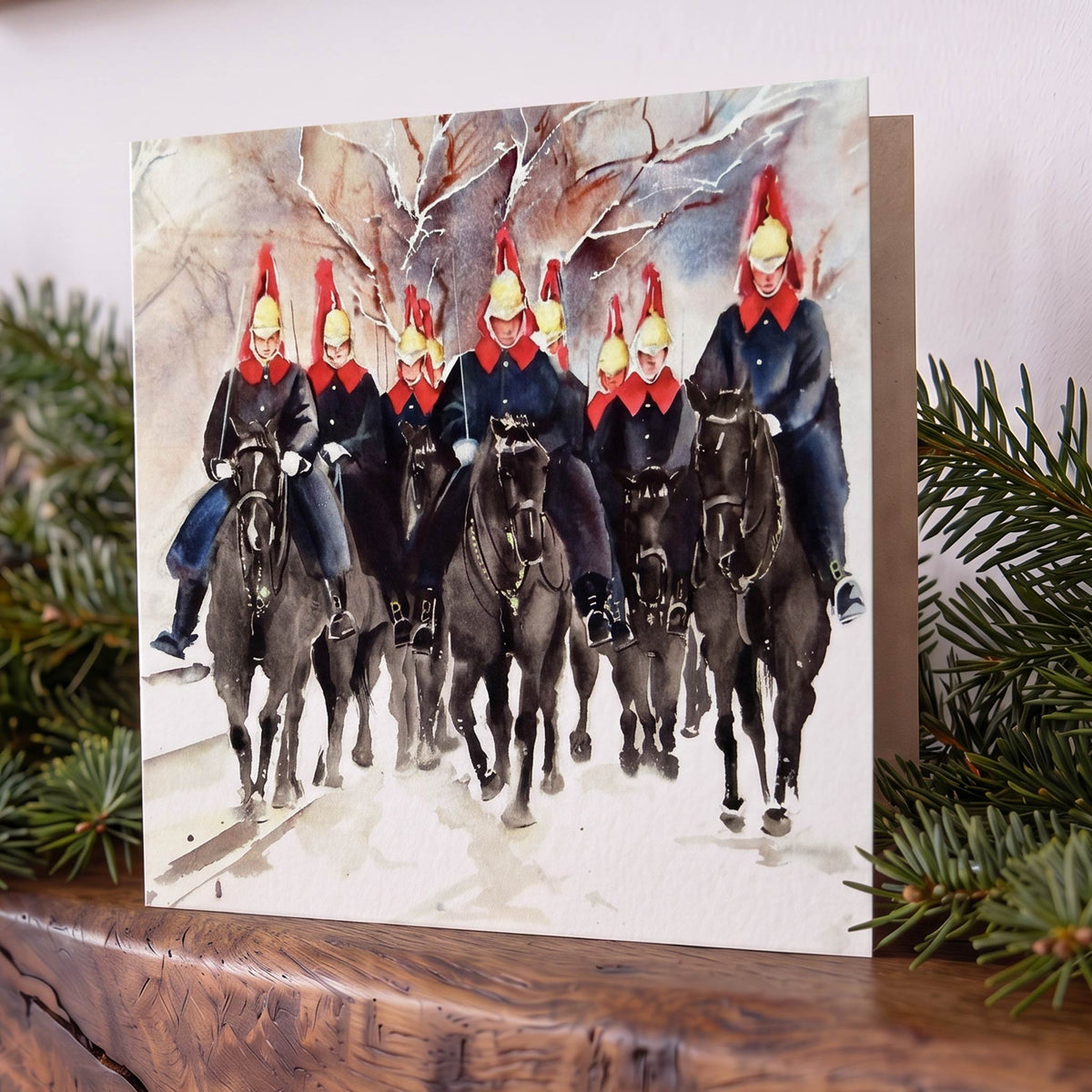 Christmas card on mantelpiece with a watercolour of Blues & Royals riders in dark uniforms, red hats and scarves crossing a snowy landscape.