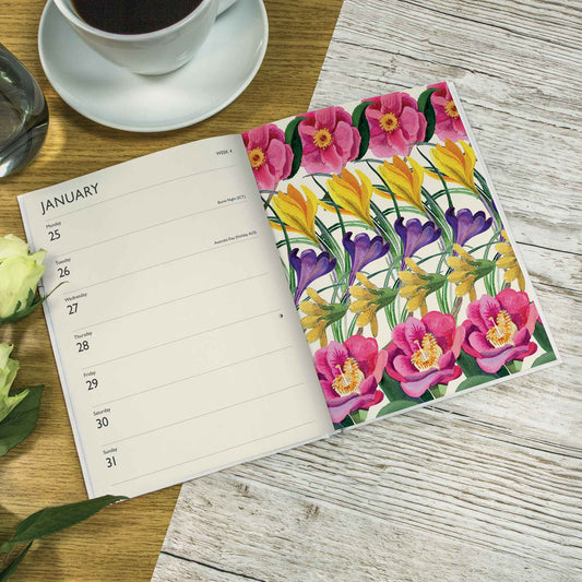 An open diary spread on a rustic wood surface, showing a floral cover design with pink tulips, yellow flowers, and purple blossoms. One page displays the January calendar layout.
