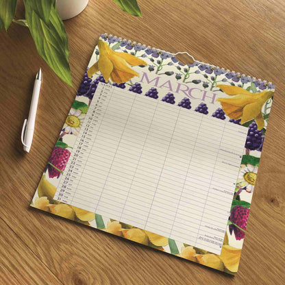 A March calendar notepad with a floral border of yellow daffodils, purple grapes, and white flowers, placed on a wooden table next to a white pen.