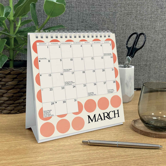 A spiral-bound desk calendar standing on a wooden table, shown in a styled office setting with a plant, scissors, a glass, and a pen. The calendar has a white cover with coral-pink circles and the word 'MARCH' printed at the bottom.