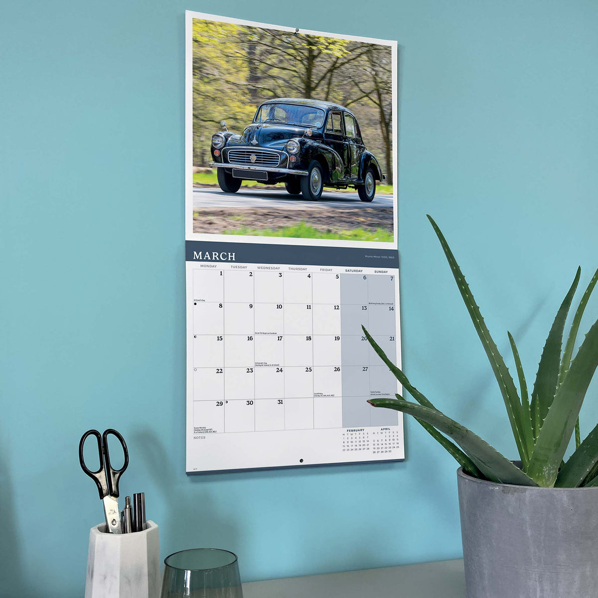 A vertical wall calendar hanging on a light blue wall, featuring a black Morris Minor 1000 photo at the top and a March calendar grid below. The calendar is styled in a home office setting with a potted aloe plant and a pencil holder on a desk.