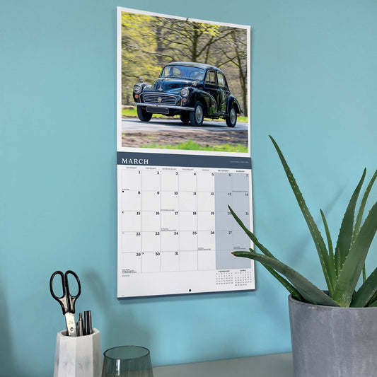 A vertical wall calendar hanging on a light blue wall, featuring a black Morris Minor 1000 photo at the top and a March calendar grid below. The calendar is styled in a home office setting with a potted aloe plant and a pencil holder on a desk.