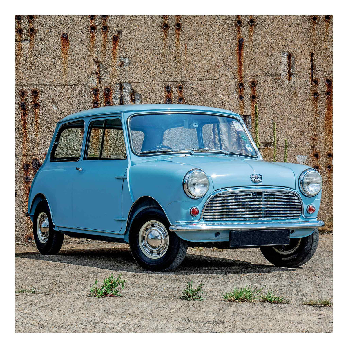 A light blue classic Mini from 1960r car parked outdoors against a weathered concrete wall with rust stains.