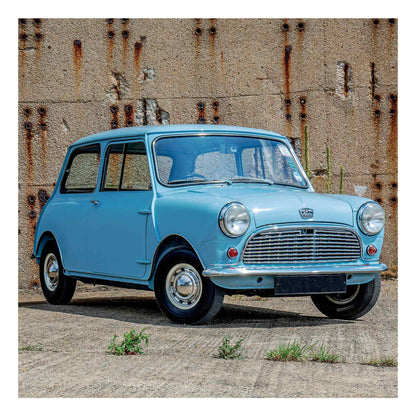 A light blue classic Mini from 1960r car parked outdoors against a weathered concrete wall with rust stains.