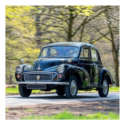 A black Morris Minor 1000 from 1969 with a chrome grille and round headlights driving on a road surrounded by blurred green trees.