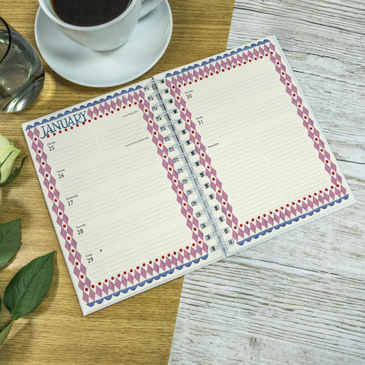 An open spiral-bound planner or agenda on a wooden tabletop, showing a January calendar page with lined daily sections and a decorative border in pink, purple, and blue. A cup of coffee on a saucer, a glass, and green leaves are partially visible around it.