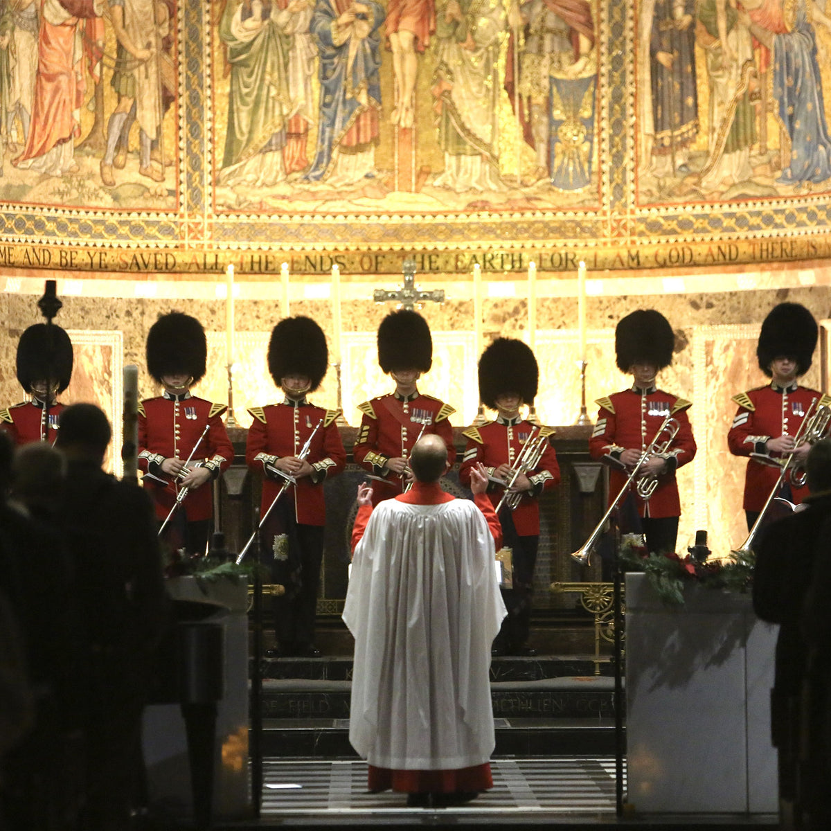 A religious service or ceremony inside a church or cathedral, with a clergyman in a white robe facing a military band of British Guards in red tunics and black bearskin hats, set against a large illuminated medieval-style mural.